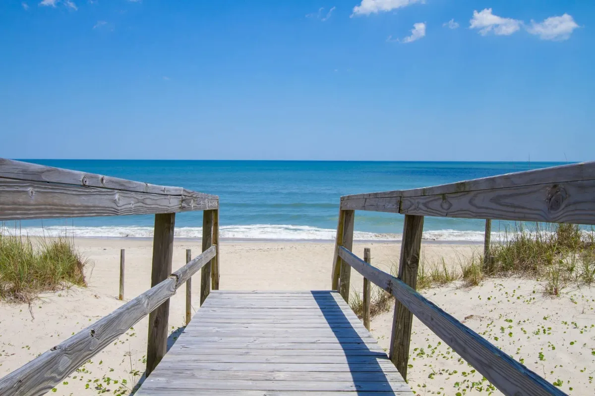 A wooden walkway leads from the property across sandy dunes to a broad, sunlit beach. The ocean is visible in the background, suggesting immediate access to the shoreline. The area around the walkway looks inviting for both people and pets to stroll and play, and the overall atmosphere is relaxed and sunny.