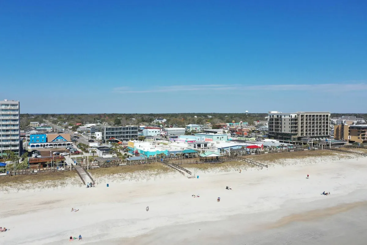 This lively exterior scene captures the vibrant Carolina Beach Boardwalk area, with colorful beachfront buildings, people enjoying the beach, and a backdrop of hotels and restaurants. The image radiates an inviting, cheerful atmosphere and showcases proximity to entertainment and dining.