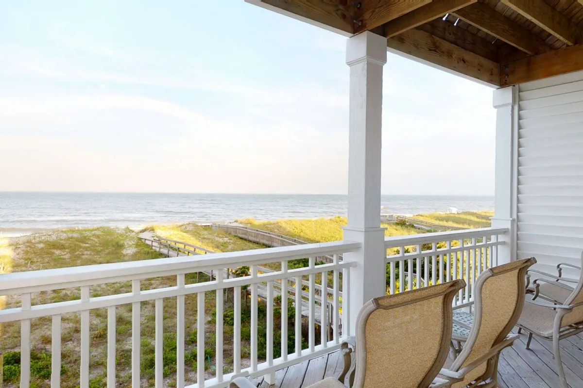 A wide oceanfront deck furnished with comfortable chairs, offering panoramic views of the beach and rolling waves. The inviting setting suggests a perfect spot for family gatherings, dolphin watching, or simply relaxing while enjoying the sea breeze. The horizon is clear, enhancing the sense of openness and connection to the water.