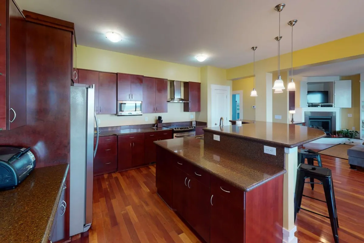 A bright kitchen scene with a central island, gleaming stainless steel appliances, wood cabinetry, and bar stools for casual dining. The layout is open and inviting, emphasizing ease of meal preparation and social interaction.