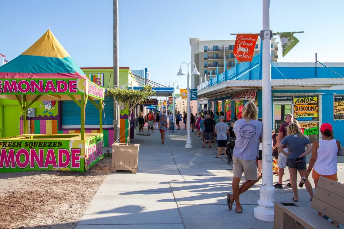 A bustling boardwalk scene with a colorful lemonade stand, bright shops, and families enjoying the festive atmosphere. The area is lively and packed with visitors, evoking the excitement of exploring local attractions, shops, and eateries near the beach.