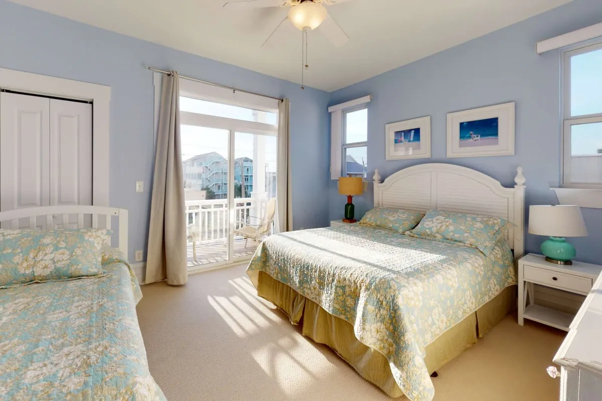 A guest bedroom filled with natural light, featuring a bed adorned with floral bedding, a decorative lamp, and framed artwork. The window offers a calming view, creating a tranquil atmosphere ideal for unwinding.