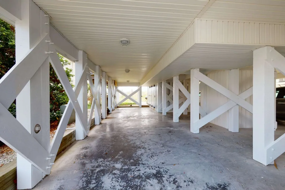 A bright, functional parking area beneath the house, featuring crisscross wooden beams, smooth concrete flooring, and subdued lighting fixtures. This spacious area offers ample room for vehicle parking and storing beach gear, highlighting the convenience for guests arriving by car.