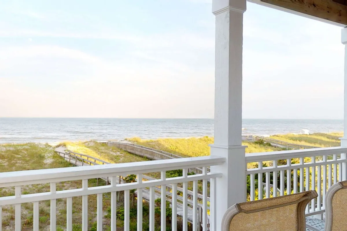 The image showcases a tranquil oceanfront scene viewed from a spacious deck, with sweeping vistas of Carolina Beach, soft beach grass lining a wooden walkway that leads directly to the sand, and the sun setting over the water. The scene is serene, inviting, and highlights the property's direct connection to the beach and the peaceful ambiance guests can experience.