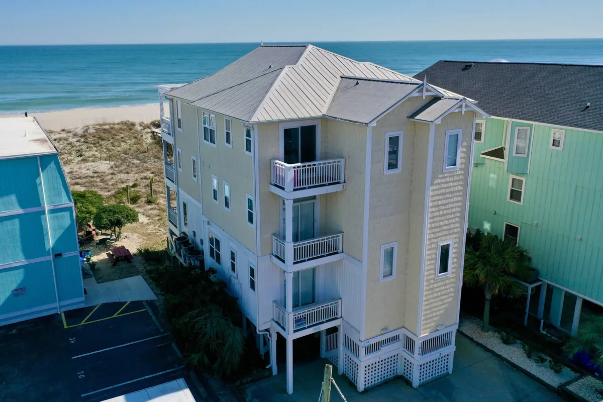 This exterior image shows a striking beachfront property with multi-level architecture and inviting balconies, surrounded by greenery and opening directly onto the beach. The property is positioned to maximize sunrise views over the ocean, with ample space for outdoor gatherings and social activities.