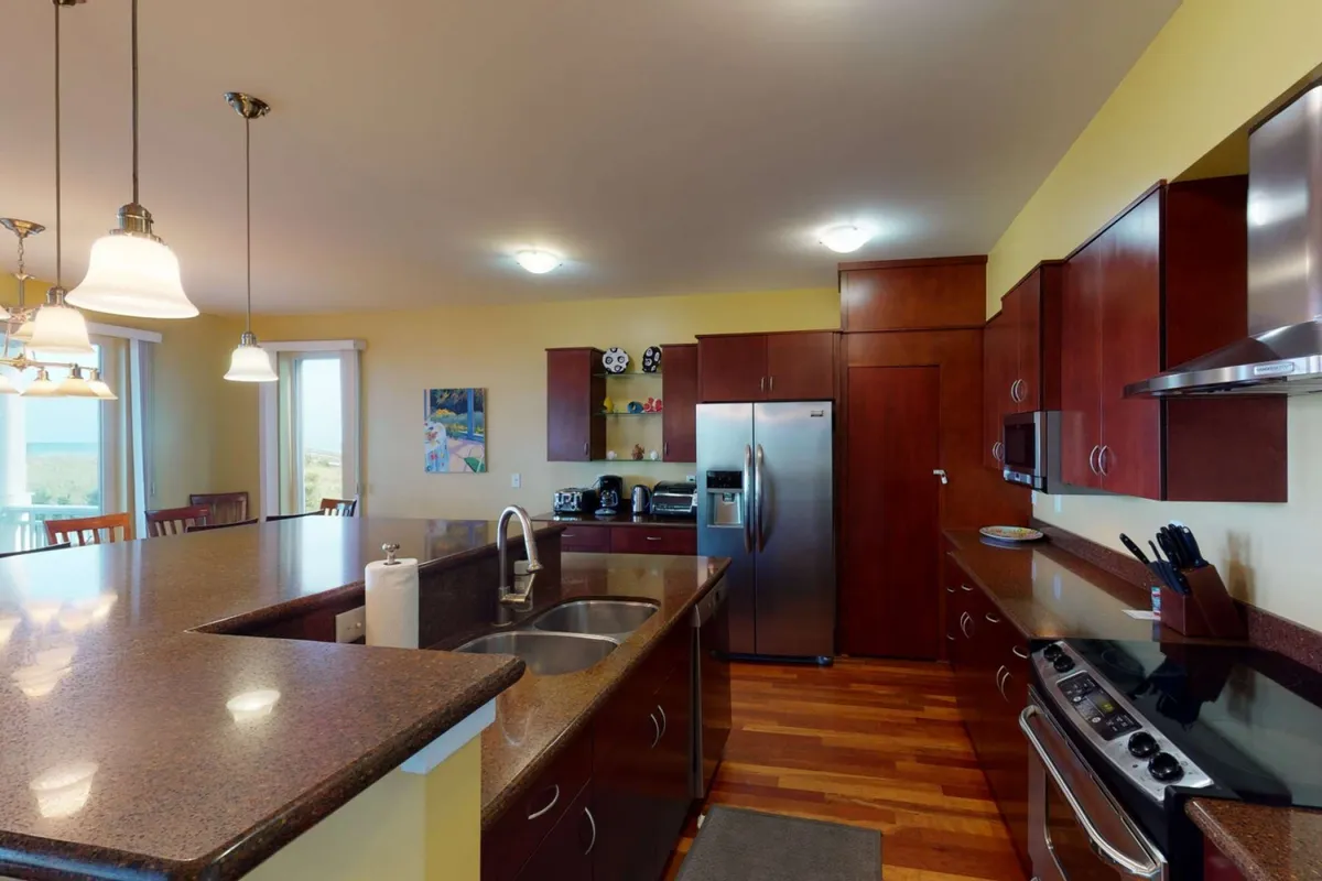 A warm, welcoming kitchen features a spacious island, stainless steel appliances including a refrigerator, oven, and dishwasher, with wood cabinetry and decorative wall art adding a homey touch. The island provides ample prep space and a gathering spot for guests to help with meal prep or socialize.