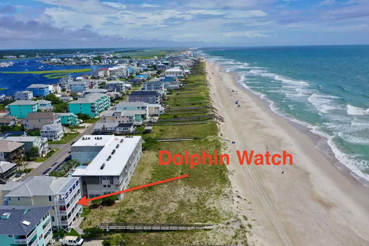 A wide-angle view of the property set against the backdrop of the beach and ocean, with the boardwalk leading directly from the home to the shore. The scene captures the vibrant coastal atmosphere and the accessibility for dolphin watching or beach activities.