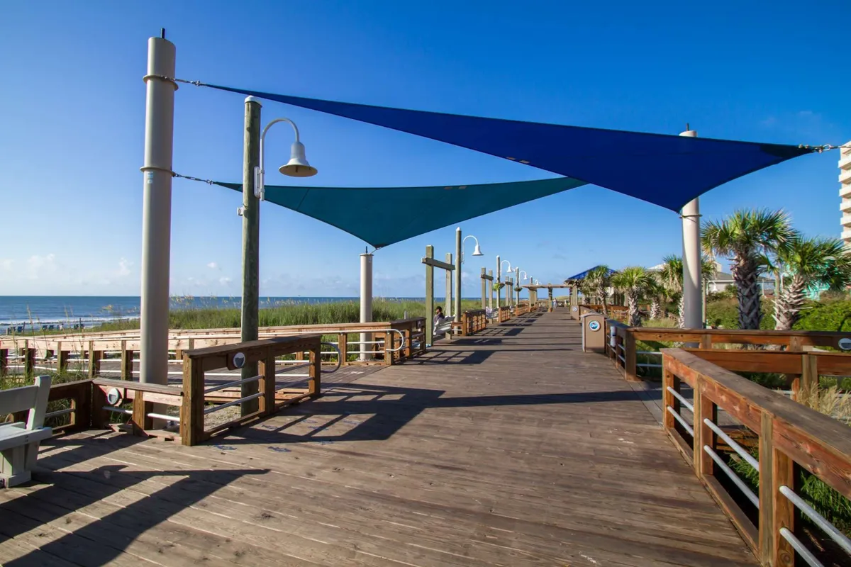A wooden boardwalk leads toward the ocean, flanked by blue shade sails, tall grasses, and lampposts. The open sky and distant water create a peaceful, inviting atmosphere that suggests direct beach access and scenic strolls.
