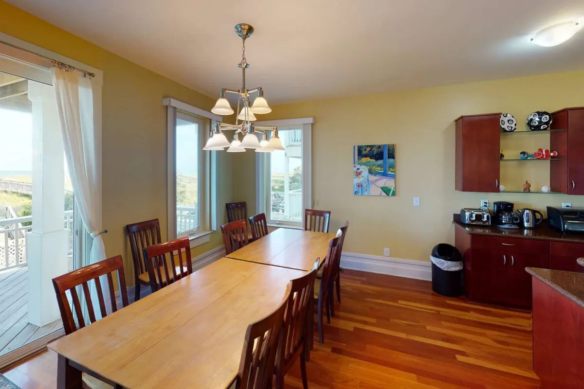A long wooden dining table set under a glowing chandelier is adjacent to the kitchen, with cabinetry and inviting décor visible in the background. The table is clearly designed to accommodate larger groups, highlighting the communal aspect of meal times.