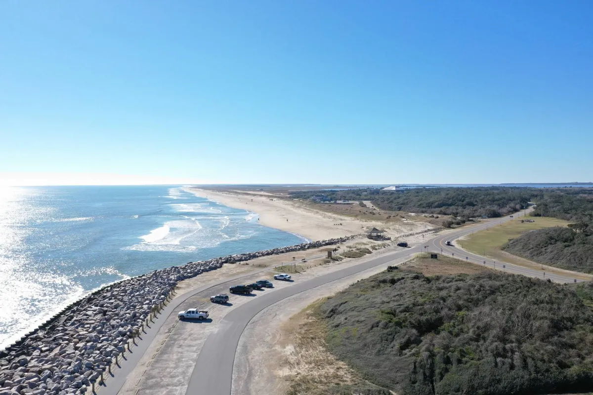 A broad view of the tranquil beach and ocean, with a road and parked cars in the foreground. The expansive shoreline is visible, with calm waves rolling in. The scene conveys a sense of openness, natural beauty, and peaceful coastal living.