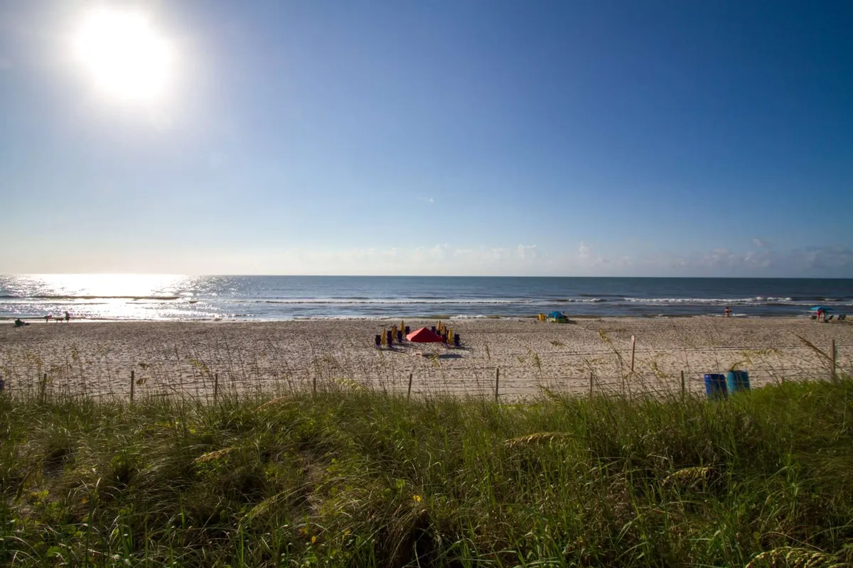 A lively scene on the beach with families and children playing in the sand, people swimming and sunbathing, and the sun shining over the ocean. The atmosphere is joyful, energetic, and filled with activity, showcasing the fun-filled beach experience accessible from the property.