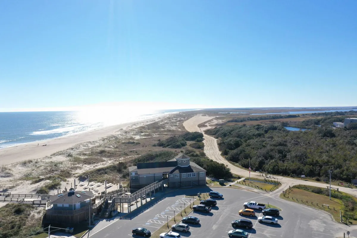 A panoramic view of a wide, sandy beach bordered by gentle ocean waves and a nearby parking area. The scene is lively but not crowded, with plenty of open space for walking, playing, and enjoying the outdoors. Several buildings in the distance hint at nearby amenities and community features.